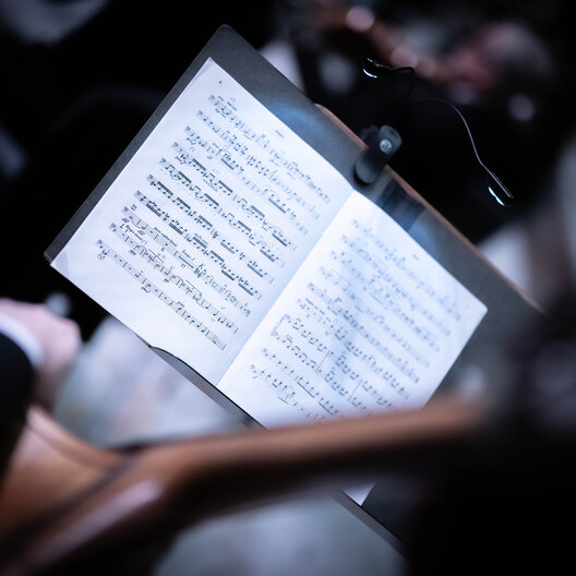 Dunkelgehaltenes Foto aus dem Orchestergraben. Im Fokus ein Notenständer mit Notenblättern, verschwommen im Hintergrund Orchestertreiben.