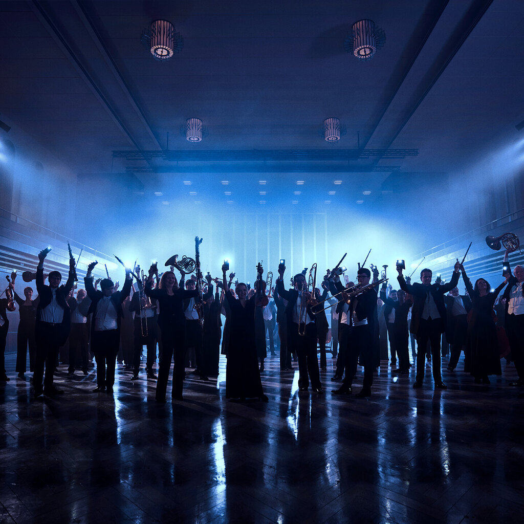 Orchester im dunklen Konzerthaus, nur Schatten der Personen sind sichtbar. Alle strecken die Hände, teilweise mit Instrumenten in der Hand, in die Luft. Orchester im dunklen Konzerthaus, nur Schatten der Personen sind sichtbar. Alle strecken die Hände, teilweise mit Instrumenten in der Hand, in die Luft. 