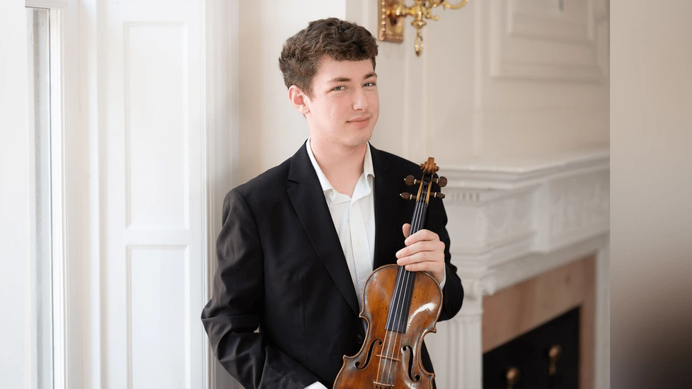 Nahaufnahme von Geiger Michael Shaham im Anzug mit Violine in der Hand in einem Raum mit weißen, verzierten Wänden. 