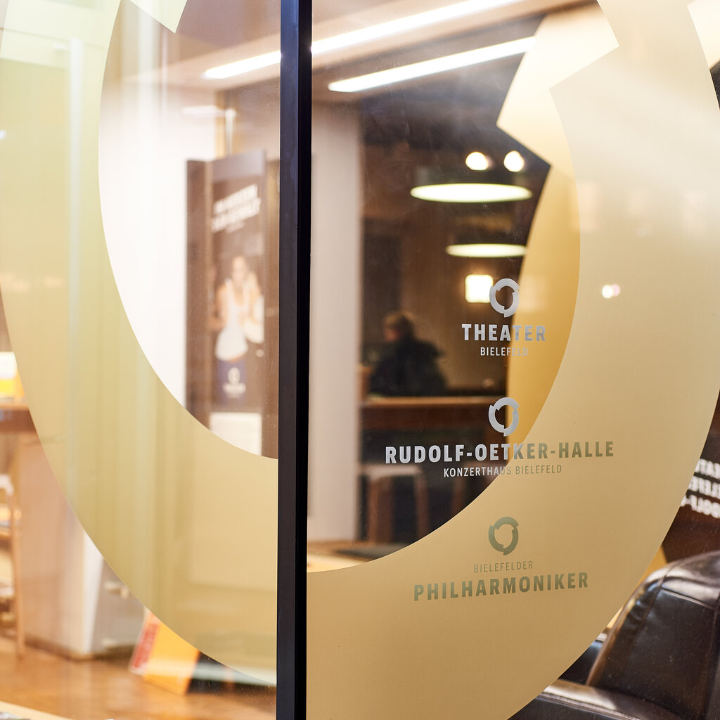 Dekoratives Bild der Fensterscheibe der Theater- und Konzertkasse. Darauf das Logo der Bühnen und Orchester Bielefeld.