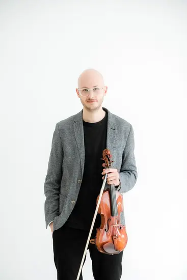 Moritz Ter-Nedden mit Violine in der Hand vor weißem Hintergrund. Er trägt eine schwarze Hose und schwarzes Oberzeil mit einer Graugemusterten Jacke, die rechte Hand in der Hosentasche.