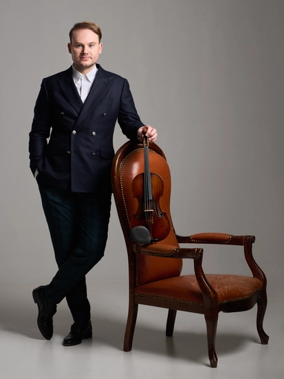 Niklas Liepe in einem Anzug stehend, mit überkreuzten Beinen und an einen Ledersessel gelehnt. Grauer Hintergrund und Boden.